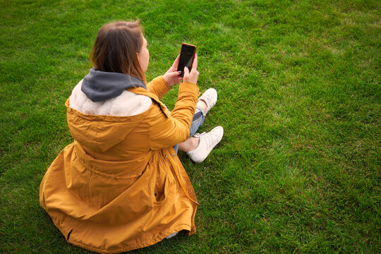 Beautiful pensive woman sits in the park on the grass and takes pictures on a smartphone