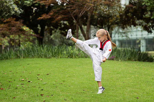 Caucasian little girl six years old in kimono with white belt exercising Taekwondo at summer park alone during coronavirus covid-19 lockdown, self isolation and social distancing