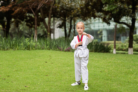 Caucasian little girl six years old in kimono with white belt exercising Taekwondo at summer park alone during coronavirus covid-19 lockdown, self isolation and social distancing