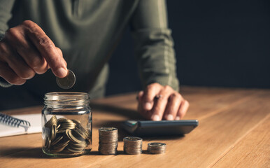 A man put coins in a glass jar and took notes to save money on a wooden table.