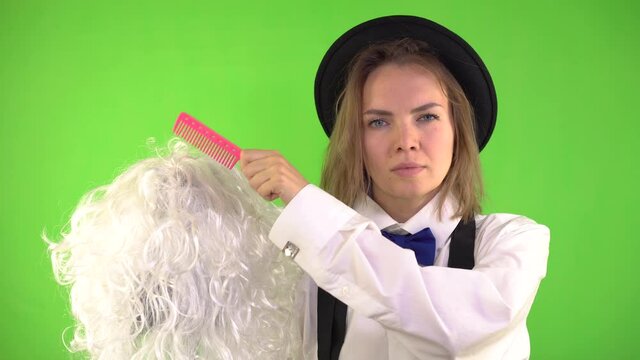 Woman in a Hat with a comb in her hand Combing a White Wig on a Green Background and Looking Seriously. Chromakey, Greenscreen. Woman combing blonde hair wig.