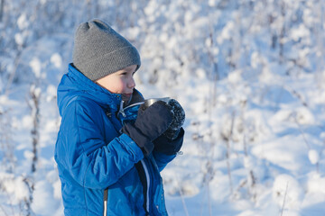 Fototapeta na wymiar Portrait of a happy boy who is drinking tea on a background of light blue landscape in winter. Concept of warming up outdoors.