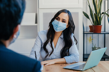 Portrait of a business woman at meeting sitting at desk with client wearing face mask for social...