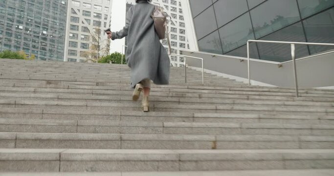 a woman is climbing the stairs,she is in a deep-gray coat and white pants,a woman looks anxious,woman carries a white bag,she walks fast.