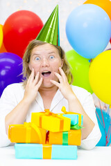 Woman with a happy smile hugging many gifts in a festive cap on a background of colorful balloons