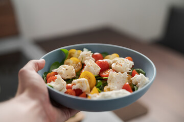 amn hand hold salad with red and yellow cherry tomatoes , mozzarella and spinach in blue bowl