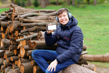Fototapeta premium teenager listening to music by headphones, relaxing in autumn city park, shows the phone screen and sitting on a log