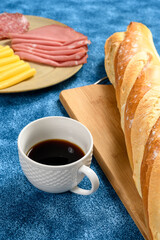 a cup of coffee with baguette italian bread dish with cheese and smoked bologna on a blue towel
