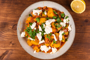 Greek vegetable stew of potatoes and vegetables in tomato sauce with chunks of soft feta cheese in a plate on a wooden table, a fork and a napkin nearby, top view
