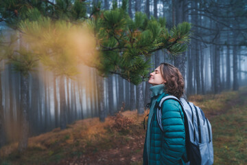 Young happy woman traveler in green jacket with backpack in foggy pine forest enjoying a walk...