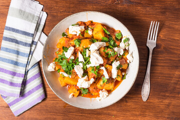 Greek vegetable stew of potatoes and vegetables in tomato sauce with chunks of soft feta cheese in a plate on a wooden table, a fork and a napkin nearby, top view