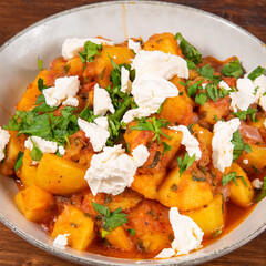 Greek vegetable stew of potatoes and vegetables in tomato sauce with chunks of soft feta cheese in a plate on a wooden table, top view, close-up