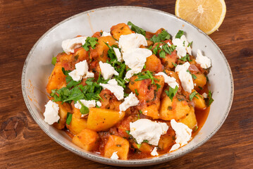 Greek vegetable stew of potatoes and vegetables in tomato sauce with chunks of soft feta cheese in a plate on a wooden table, top view, close-up