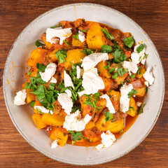 Greek vegetable stew of potatoes and vegetables in tomato sauce with chunks of soft feta cheese in a plate on a wooden table, top view, close-up