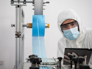 The engineer worker in protective suits, masks, and gloves is checking and controlling the machine in the manufacture of surgical masks for coronavirus..