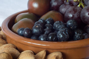 Close Up of some blueberries with a bunch of black grapes, tomatoes, and almonds in a clay bowl on a kitchen table in a home. Nuts, vegetables, and fruit. Healthy food.