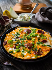 Delicious breakfast - scrambled eggs with fried sausages, cottage cheese and vegetables served on black plate on wooden table
