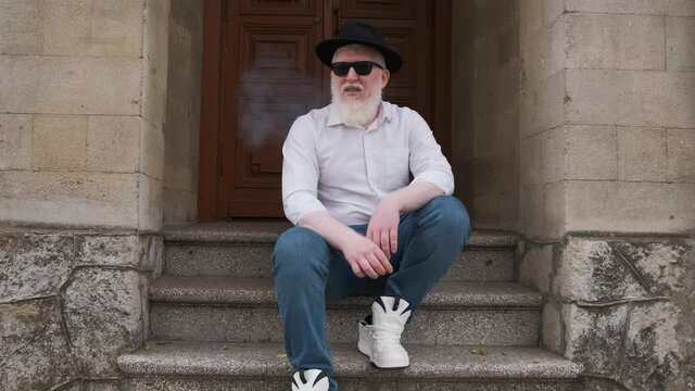 Man suffering from albinism sits outside on the steps and smokes a cigarette while wearing a hat and glasses to protect himself from the sun.