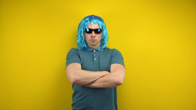 A funny young man in a woman's blue wig and sunglasses crossed his arms seriously. Shooting in the studio on a yellow background.