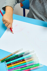 Cute little boy with blond hair draws colored pencils at home. Draws at the blue table. Close up of child's hands drawing at white paper