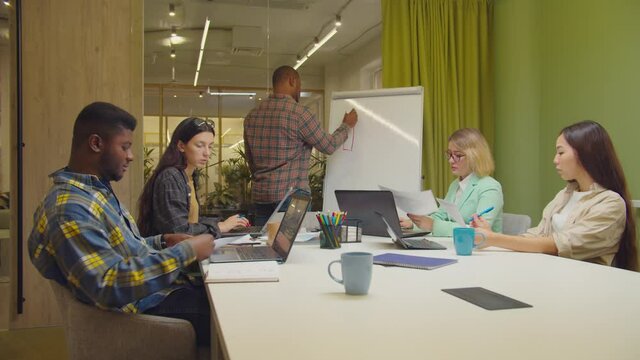 Confident african american male manager drawing financial graph on flipchart, pointing and setting business goals for diverse multiracial business team while working together on project at office.