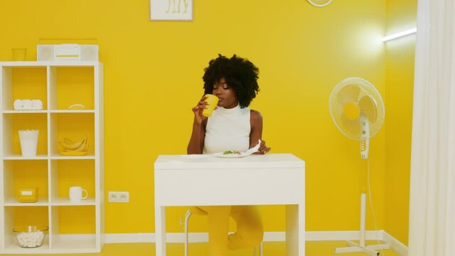 Beautiful black woman is sitting at white desk in creative yellow office, having snack time, eating pizza with cheese and drink beverage, art video concept styled in yellow and white, Slow motion.