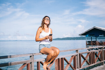 Happy hipster tourist in sunglasses resting at nature environment with seashore on background, cheerful woman with cellphone enjoying summer vacations for international visiting and solo travelling
