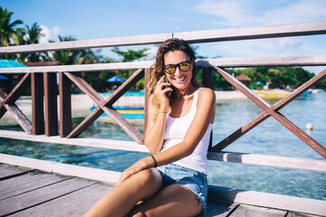 Cheerful wanderer in sunglasses resting at sea pier and calling to best friend for discussing getaway solo vacations in Indonesia, happy woman enjoying recreation time in tropical resort paradise