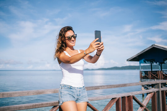 Cheerful millennial girl in stylish sunglasses using mobile and making selfie pictures and share positive content to blog followers, happy female tourist enjoying summer vacations for visiting Maldive