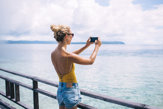 Millennial female tourist using cellphone camera for photographing sea landscape of Bahamas enjoying summer vacations, woman with smartphone device making picturesque images of Hawaii scenery