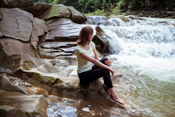 Young Woman is sitting at stone Looking at Splashing Waterfall. Peaceful Caucasian Traveler Sit at Beautiful Stream.