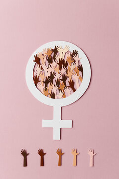 Multi racial hands filling a venus symbol cut out of paper on pale pink background