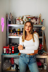 Stylish artist in casual clothes posing for the camera in the workshop on the background of shelves with painting tools. Portrait of a sexy artist in the studio for painting