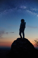Full length of brave cosmonaut standing on rocky mountain with breathtaking starry sky, Milky way on background. Silhouette of space traveler wearing space suit with helmet. Concept of space traveling