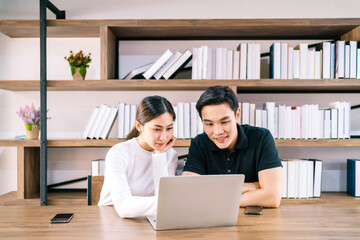 Young Asian male and female working together on the wooden table with laptop, looking at the laptop and smiles with a bookshelf in the background.