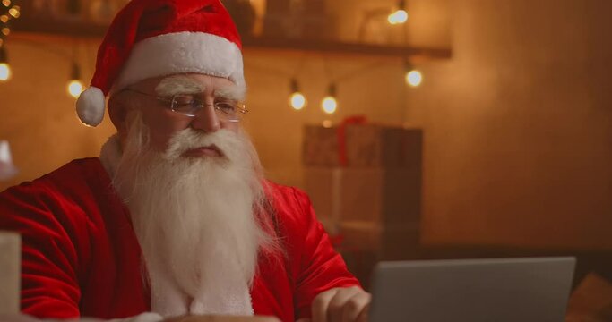 Real Santa Claus using new technology for communication with children, receiving mail or wish list. Cheerful working on laptop and smiling while sitting at his chair with fireplace and Christmas Tree