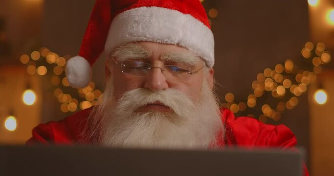 Real Santa Claus using new technology for communication with children, receiving mail or wish list. Cheerful working on laptop and smiling while sitting at his chair with fireplace and Christmas Tree