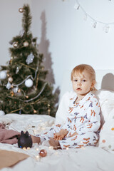 the child's first Christmas. a pensive little boy is lying in his holiday pyjamas on a bed against the background of a brightly decorated fir tree with a Golden light garland.