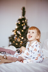 Obraz na płótnie Canvas the child's first Christmas. a pensive little boy is lying in his holiday pyjamas on a bed against the background of a brightly decorated fir tree with a Golden light garland.