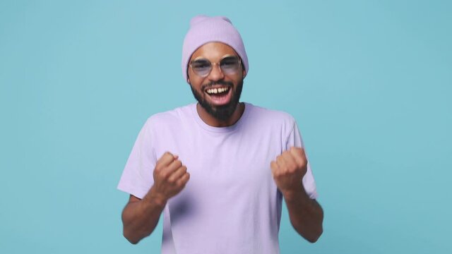 Happy joyful young african american man 20s wearing violet t-shirt hat glasses isolated on blue background in studio. People lifestyle concept. Doing winner gesture clenching fists say yes dancing