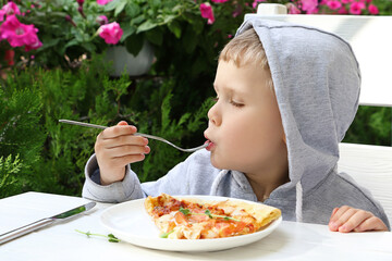 Pretty toddler is holding fork with a slice of pizza.The look of pleasure on his face.Beautiful floowers and summer foliage n the backgrund.Open cafe interior.