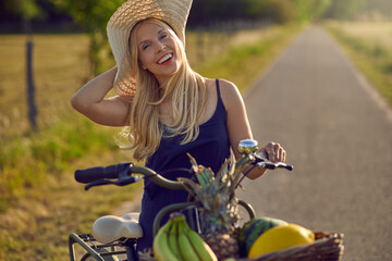 Happy laughing carefree middle-aged blond woman standing on a rural road with a basket of fresh vegetables and fruit on her handlebars of her bicycle on a sunny summer day