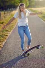 Happy smiling agile middle-aged blond woman playing on her skateboard in warm evening light on a narrow country lane between green fields