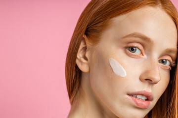 portrait of young woman with cream on cheek. Attractive caucasian woman with red hair, clean fresh freckled skin, posing against pink wall. Natural beauty concept.