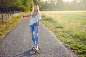 Fit active middle-aged woman playing on her skateboard approaching the camera along a narrow rural road with a happy smile backlit by the evening sun