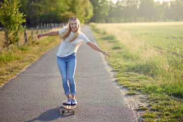 Fit active middle-aged woman playing on her skateboard approaching the camera along a narrow rural road with a happy smile backlit by the evening sun