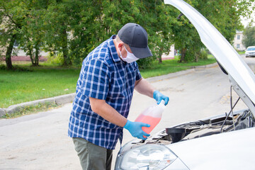 a man in a blue checkered shirt and a blue medical mask is standing by the car with the hood open, he opens the canister to top up the liquid in the car.
