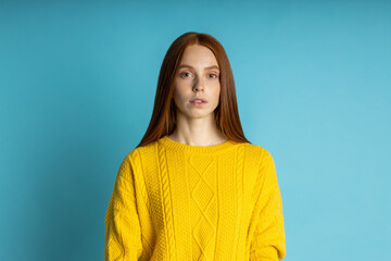 Fototapeta premium redhead beautiful caucasian young woman with healthy freckled clean skin, in yellow sweater looking at camera with calm and serious expression om blue background. Closeup studio shot