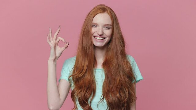 Smiling amazed ginger redhead young woman in casual blue turquoise t-shirt posing isolated on pink color background studio. People lifestyle concept. Looking camera say wow showing OK gesture blinking