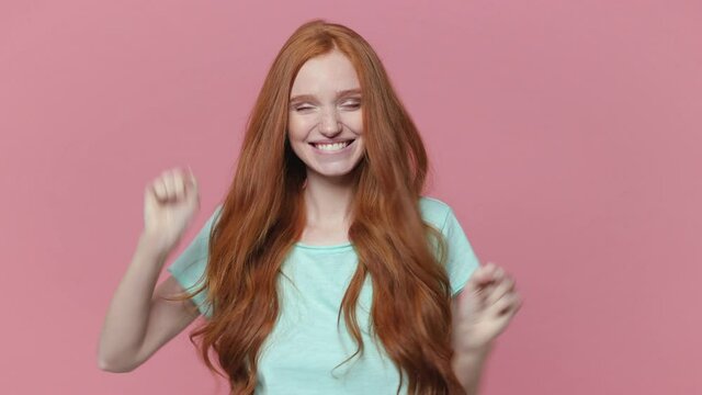Joyful excited redhead young woman in blue turquoise t-shirt posing isolated on pink background in studio. People lifestyle concept. Looking camera doing winner gesture clenching fists jumping say yes